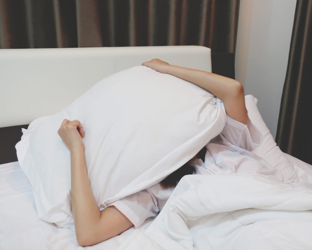 Person lying in bed, covering head with a pillow and blanket, with arm visible. Room features a white headboard and curtains in the background.