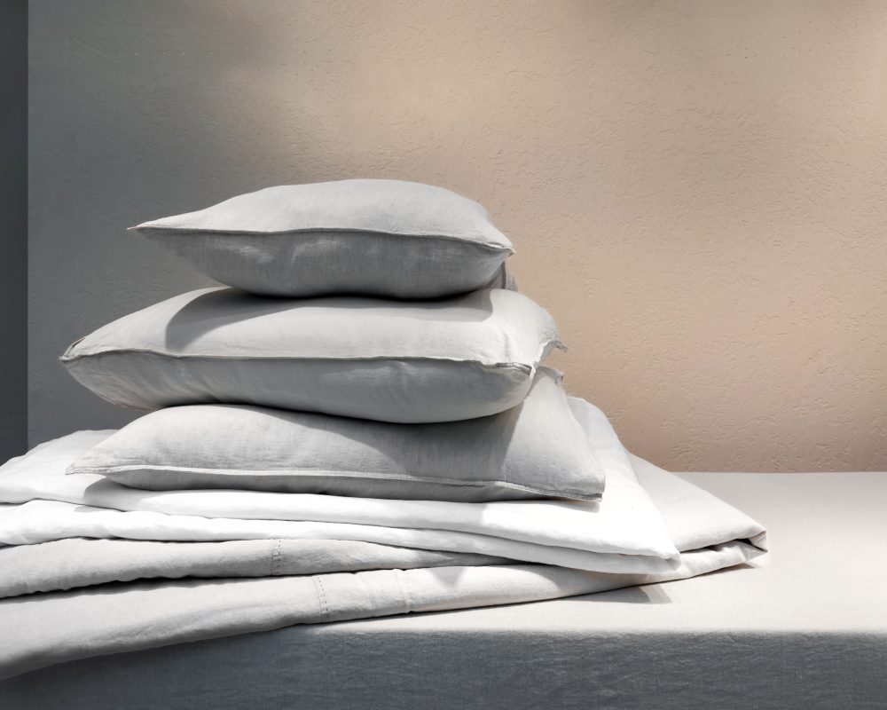 A stack of white pillows and folded bedsheets on a white table with a neutral background.