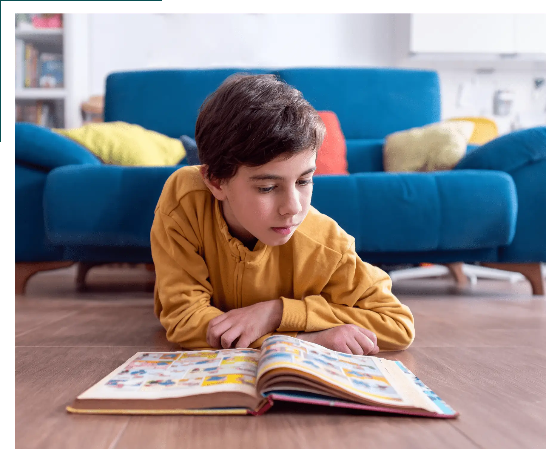 A boy in a yellow sweater lies on the floor reading a book in a living room with a blue sofa and colorful pillows in the background.